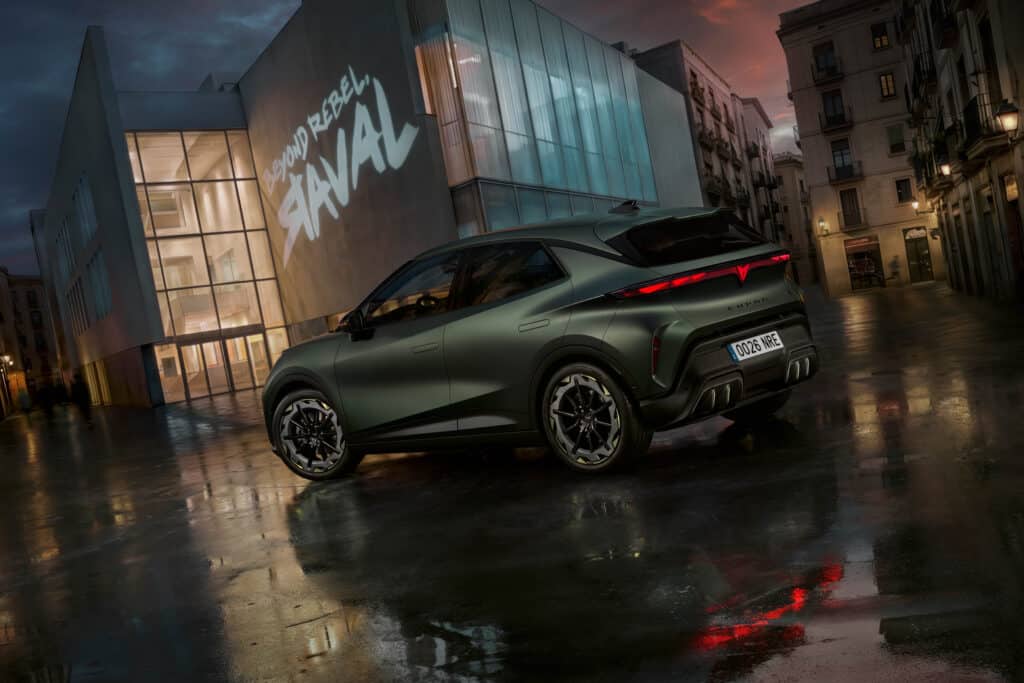 Auf diesem Bild ist ein grünes Cupra Raval Concept Car auf einer nassen städtischen Straße bei Dämmerung zu sehen, umgeben von einem modernen Gebäude mit der projizierten Aufschrift 'BEYOND REBEL, RAVAL' und traditionellen Stadthäusern.